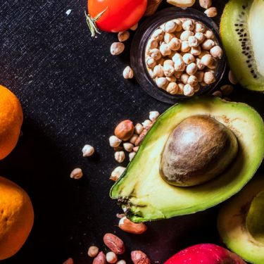 A vibrant arrangement of fruits and nuts on a black background featuring kiwi, avocado, oranges, apple, and peanuts.